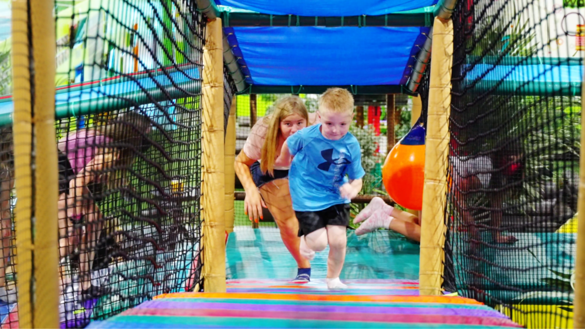 Untitled design (22) Two happy children enjoying a vibrant indoor playground, sliding down a bright red slide surrounded by colorful play structures and playful decor.